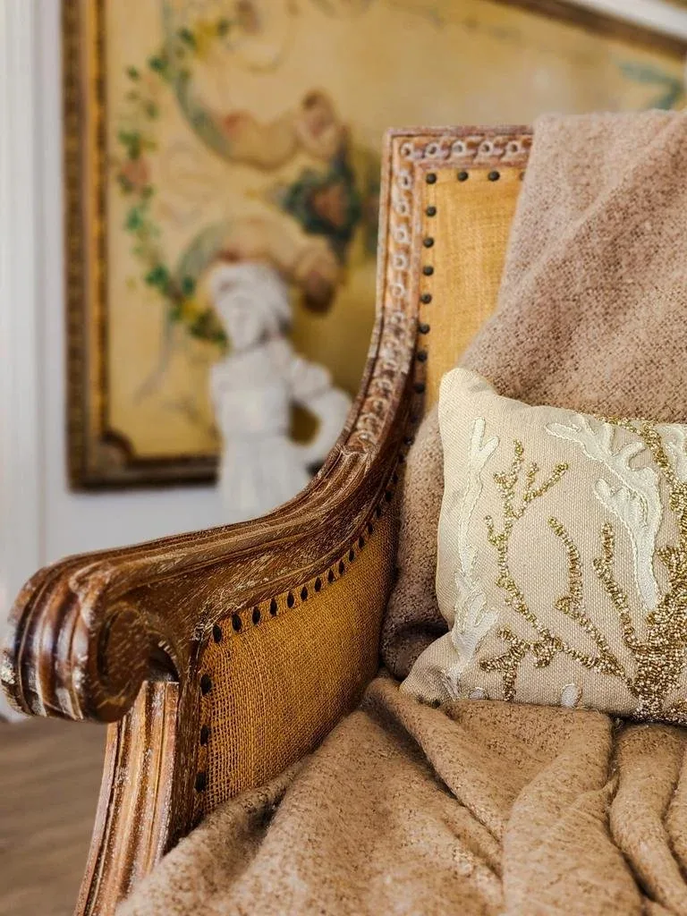 Ornate carved armchair layered with a cashmere throw and gold embroidered pillow beneath tapestry art.