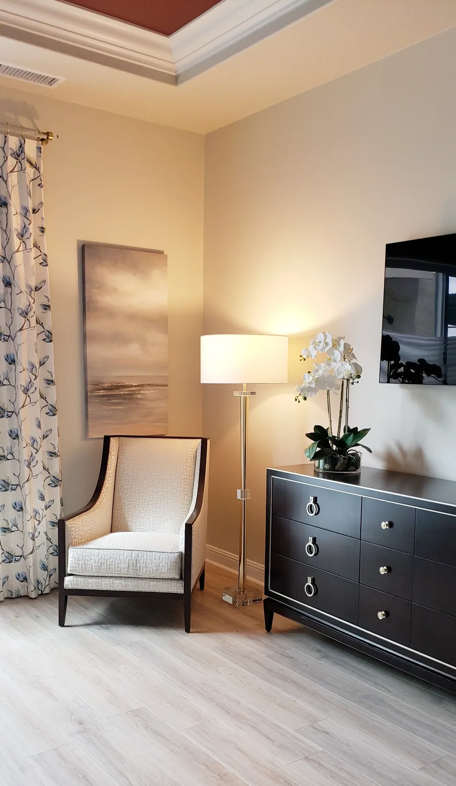 Bedroom seating nook with an upholstered chair, dark dresser, table lamp and white orchid.